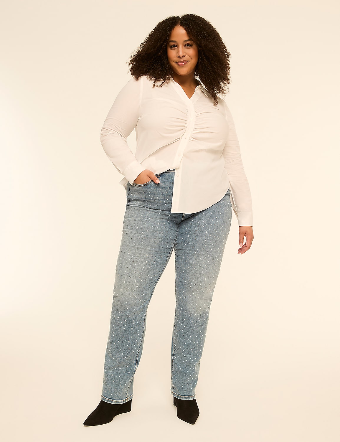 A woman with curly hair stands in the foreground wearing a white ruched shirt and embellished jeans, against a plain, light-colored background. Product Image 3
