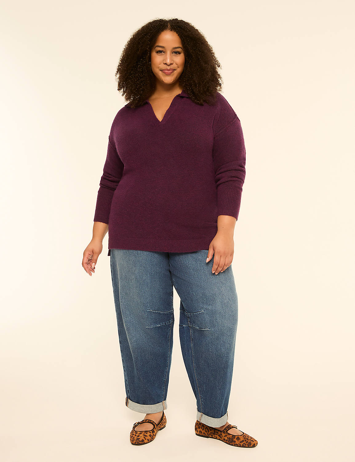 A woman with curly dark hair stands in the foreground wearing a plum-colored V-neck sweater and baggy denim jeans, against a plain cream-colored background. Product Image 3