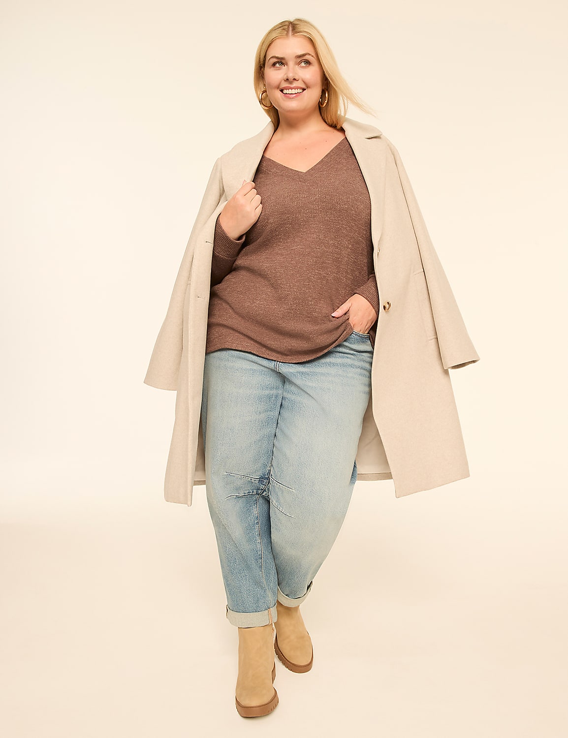 A smiling woman in a brown sweater, light blue jeans, and tan boots stands against a plain, light-colored background with a cream coat draped over her shoulders. Product Image 3