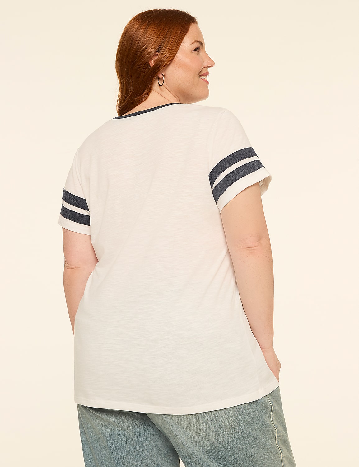 A woman with red hair wearing a white t-shirt with navy stripes on the sleeves and light blue jeans stands against a plain, light-colored background. Product Image 2