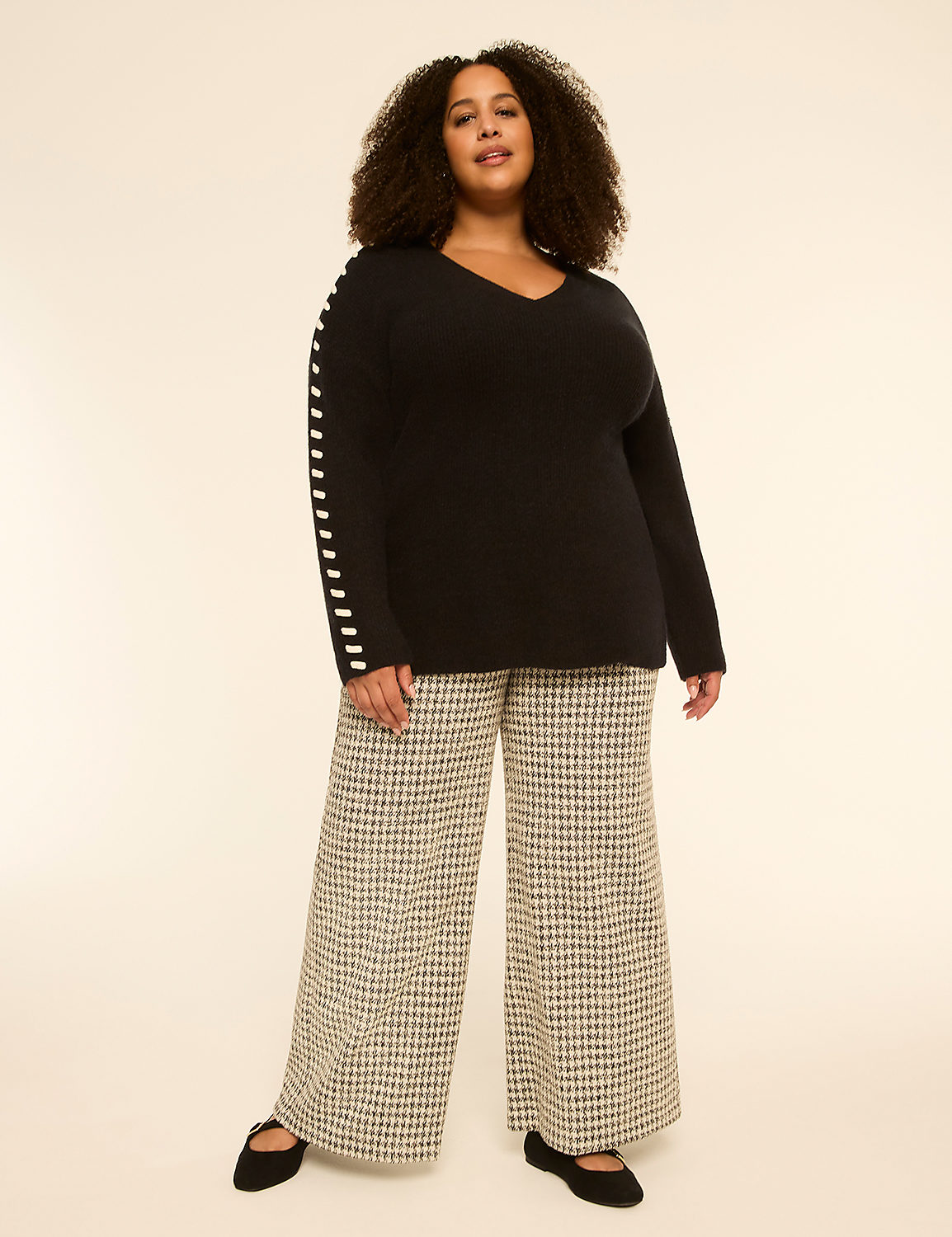A woman with curly hair stands in a black v-neck sweater with striped sleeves and wide-leg checkered pants against a plain, light-colored background. Product Image 3