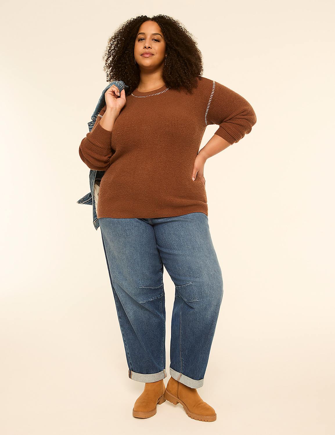 A woman in a brown sweater and jeans stands against a plain, light-colored background, with a denim jacket slung over her shoulder. Product Image 3