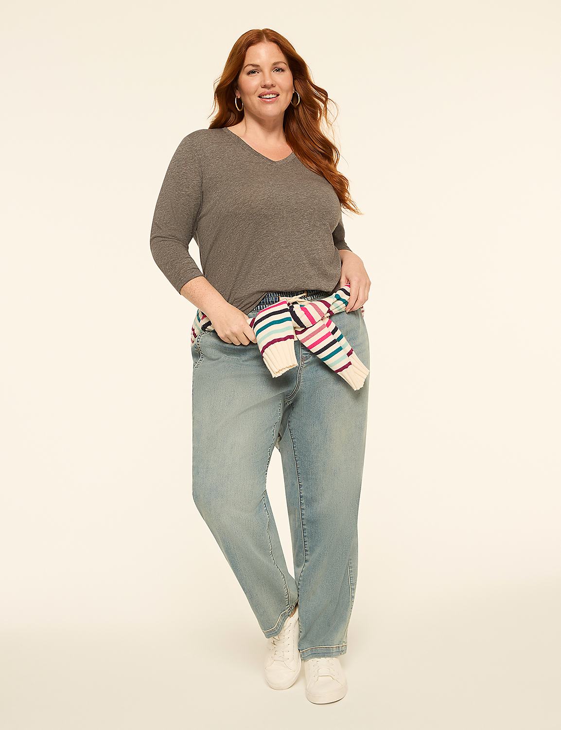 A woman with red hair stands in the foreground wearing a gray v-neck shirt, light wash jeans, and white sneakers, with a striped sweater tied around her waist, against a plain, light-colored background. Product Image 3