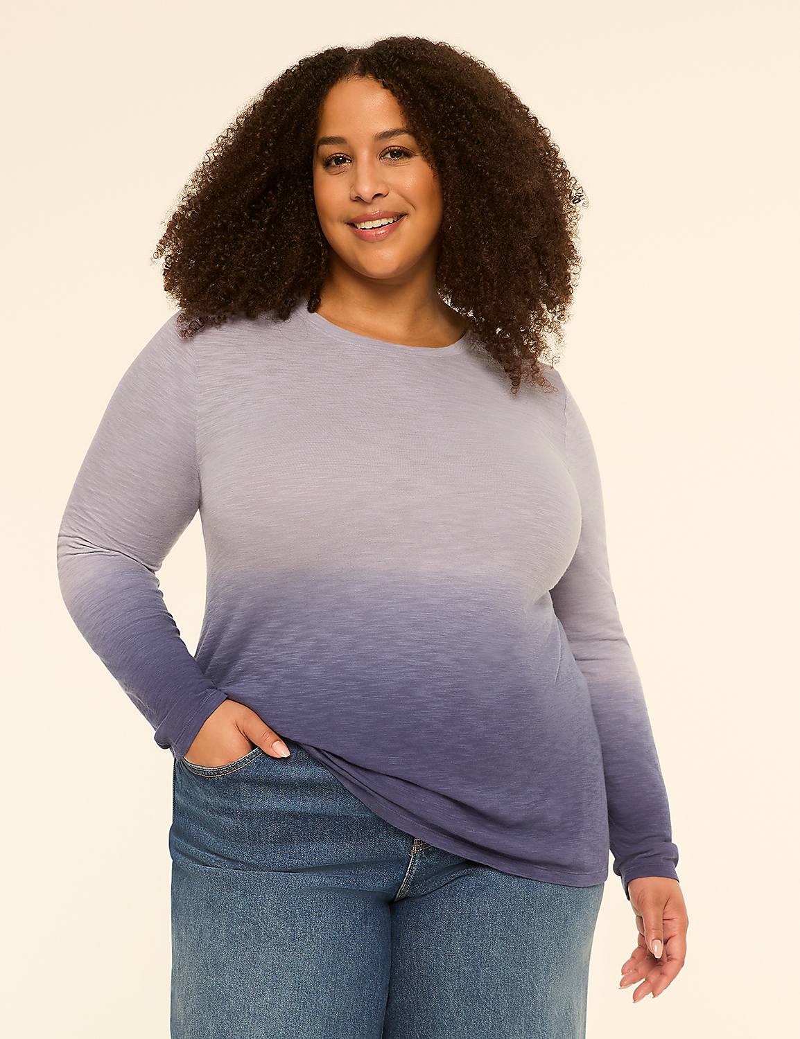 A smiling woman with curly hair in a blue ombre long-sleeved shirt and jeans stands against a plain cream background. Product Image 1