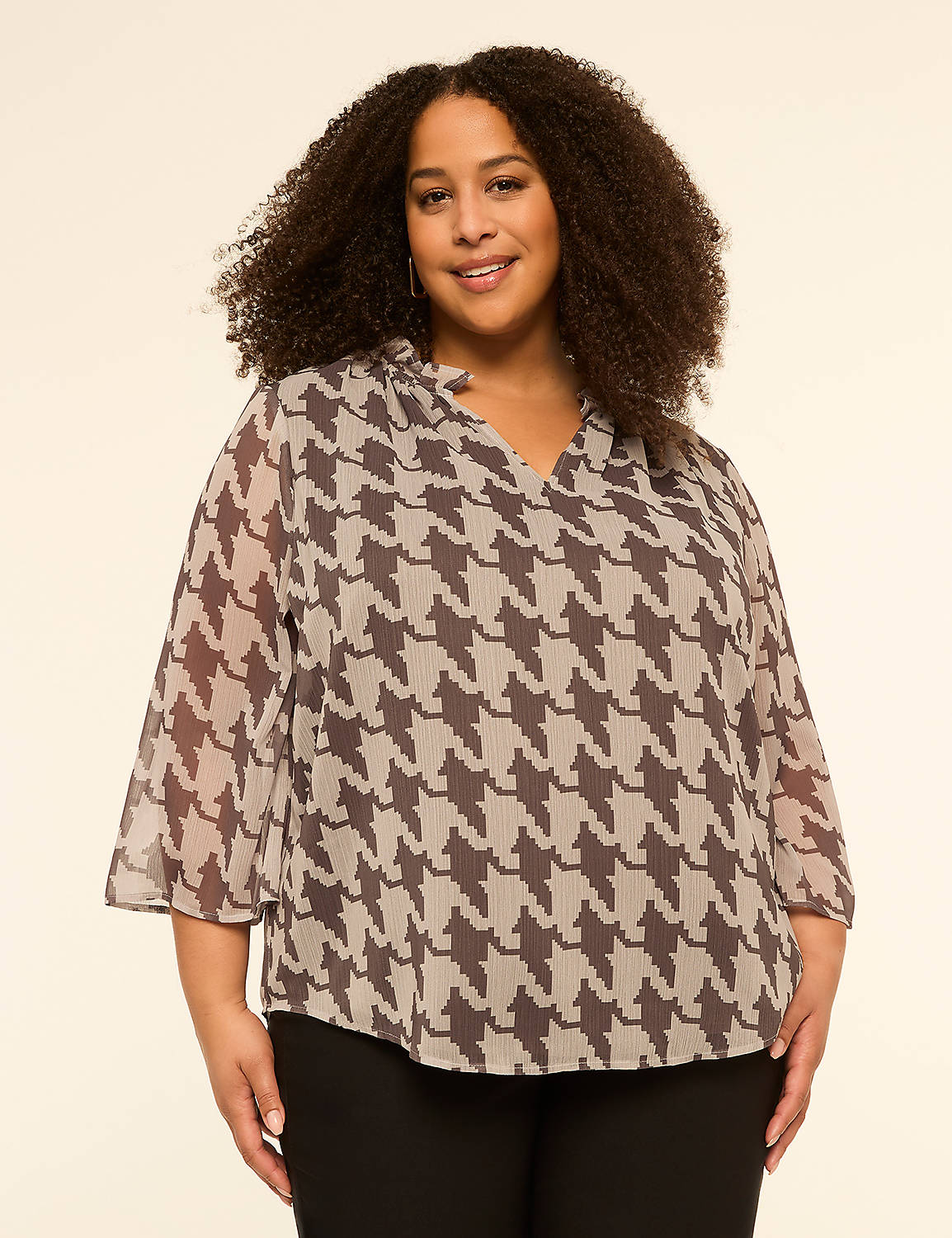 A woman with curly hair smiles at the camera, wearing a patterned blouse and black pants against a plain, light-colored background. Product Image 1