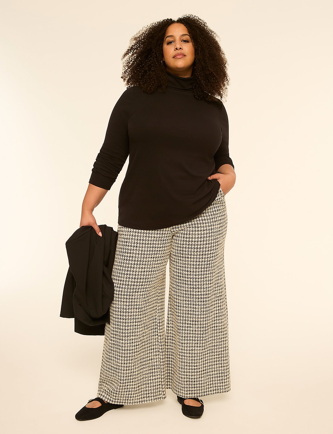 A woman with curly hair stands in the foreground wearing a black turtleneck and patterned wide-leg pants, holding a black jacket, against a plain, light-colored background. Product Image 3