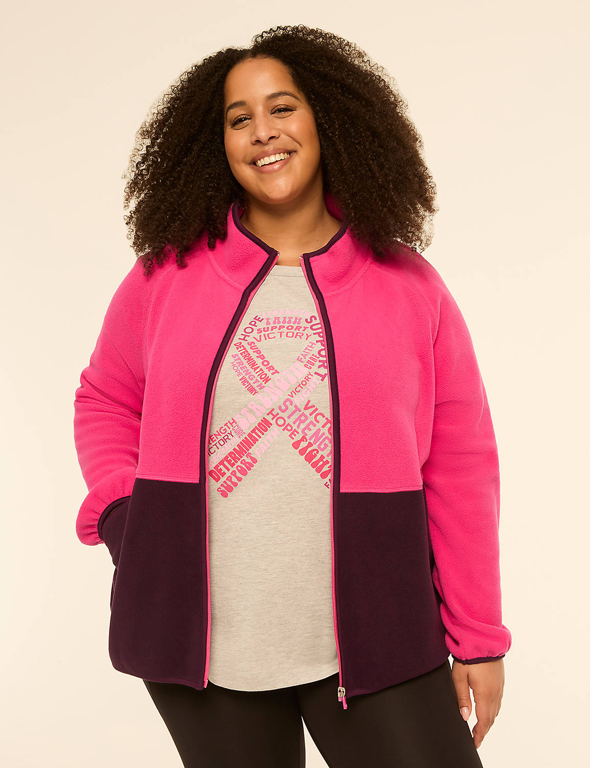 A smiling woman with curly hair stands in the foreground wearing a pink and purple zip-up jacket over a grey t-shirt with a pink ribbon design, against a plain, light-colored background. Product Image 1