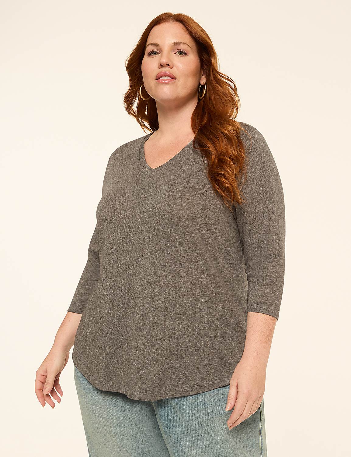 A woman with red hair and hoop earrings poses in a grey v-neck shirt and light blue jeans against a plain, light background. Product Image 1
