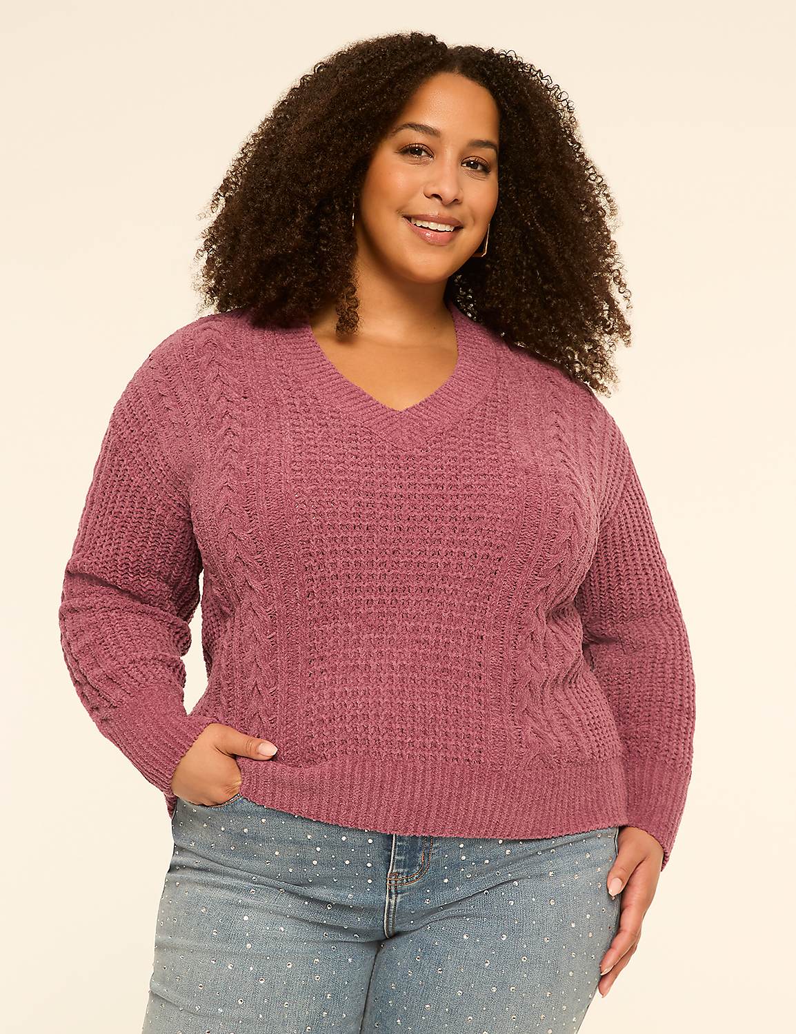 A smiling woman with curly hair in a mauve cable-knit sweater and polka-dot jeans stands against a plain cream background. Product Image 1