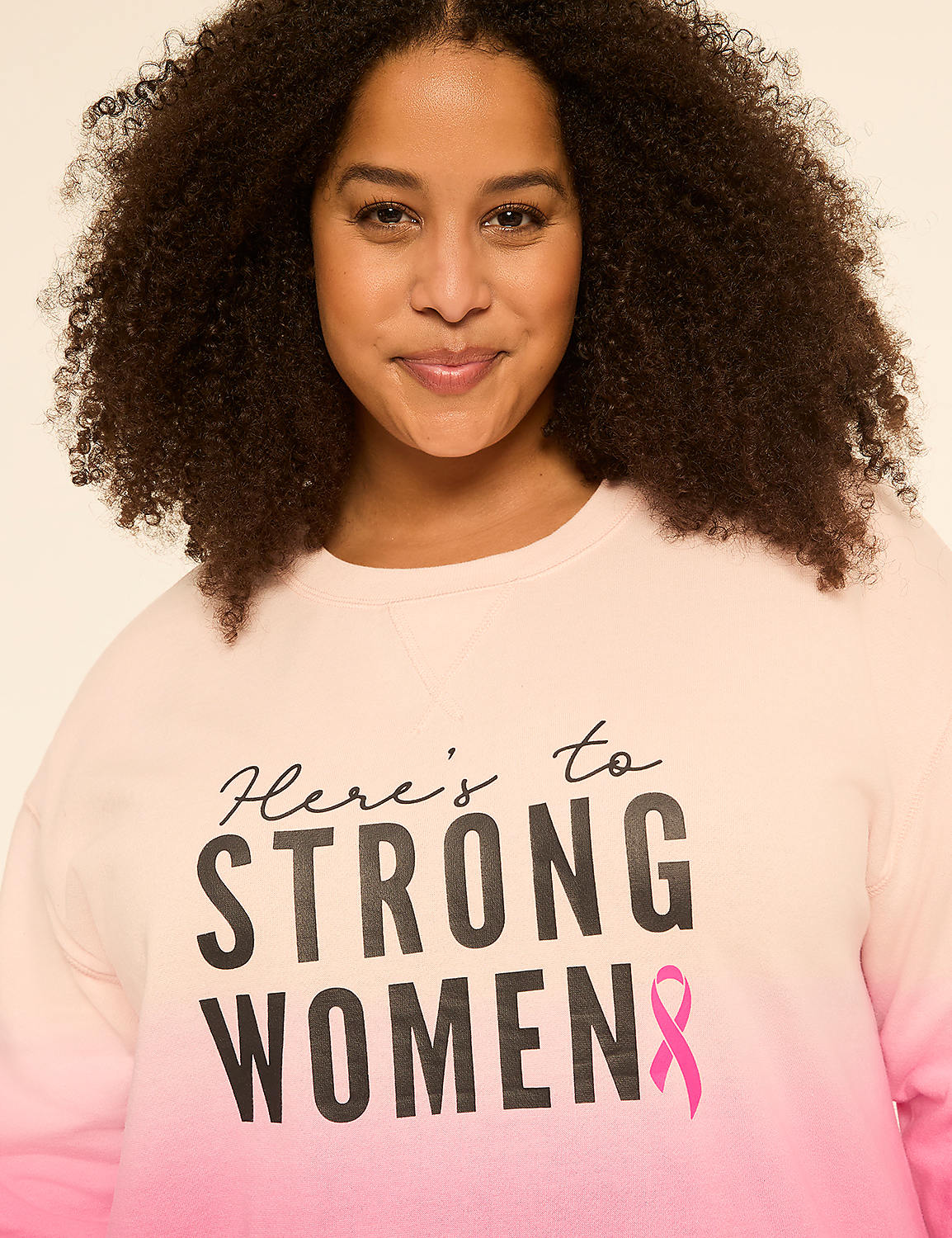 A woman with curly hair smiles at the camera, wearing a pink and white ombre sweatshirt with the words "Here's to STRONG WOMEN" printed on it, against a plain, light background. Product Image 4
