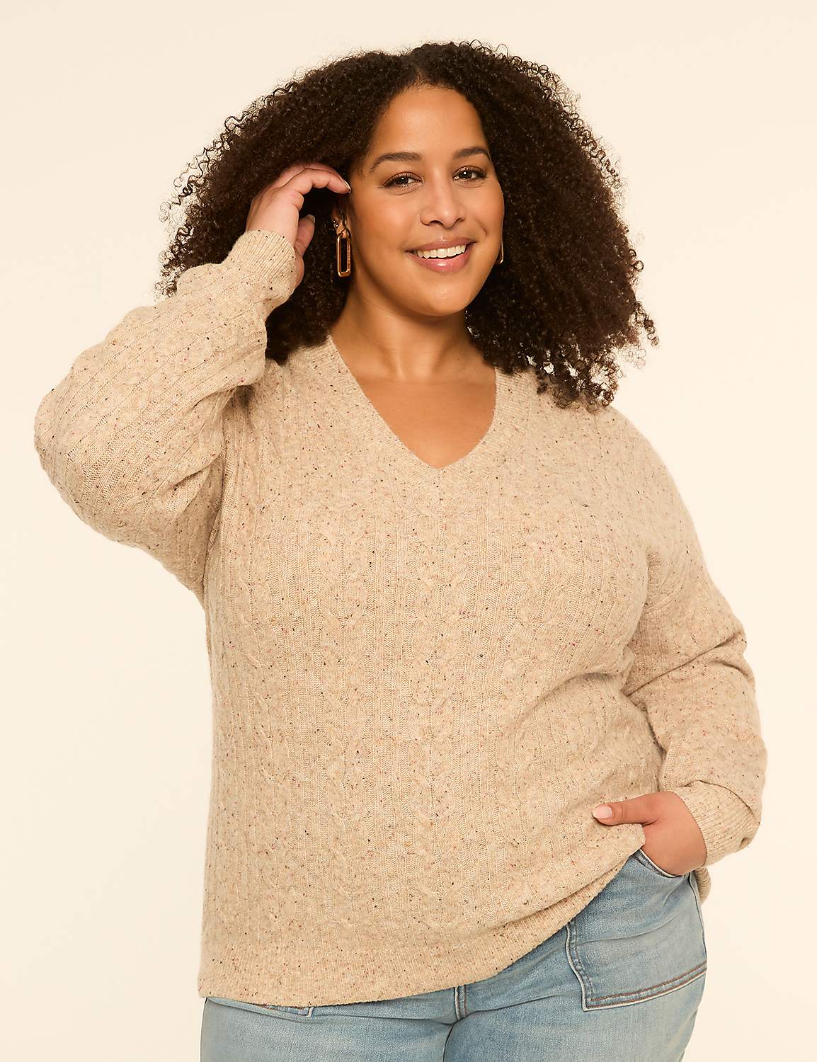 A smiling woman with curly hair in a beige sweater and jeans stands against a plain, light-colored background. Product Image 1