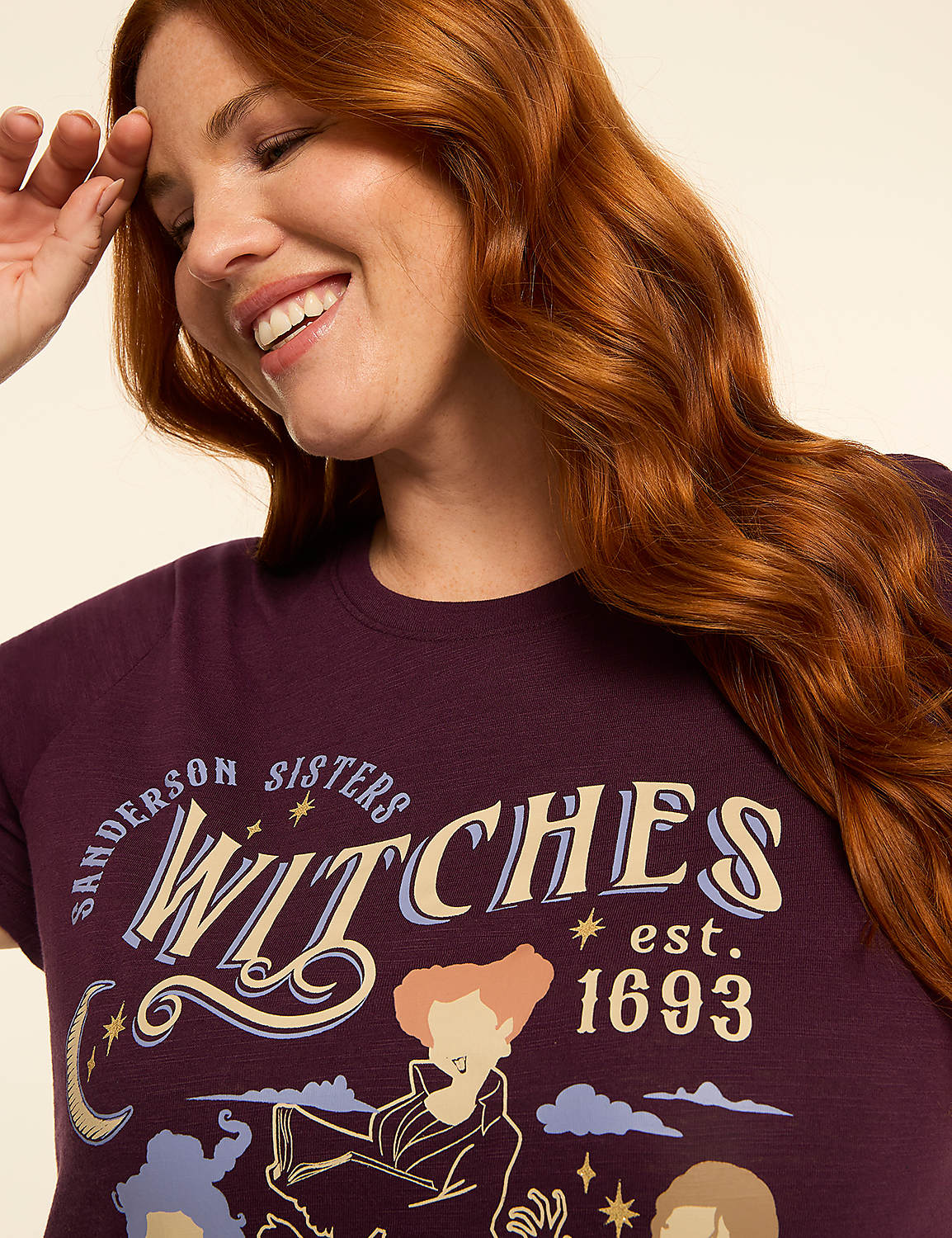 A smiling woman with auburn hair is wearing a dark purple t-shirt with a graphic of the Sanderson Sisters from Hocus Pocus, set against a plain, light-colored background. Product Image 4