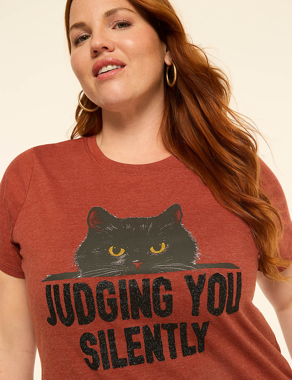 A woman with auburn hair and hoop earrings smiles slightly in the foreground, wearing a rust-colored t-shirt with a black cat graphic and the words "JUDGING YOU SILENTLY" printed below it, against a plain, light-colored background. Product Image 4