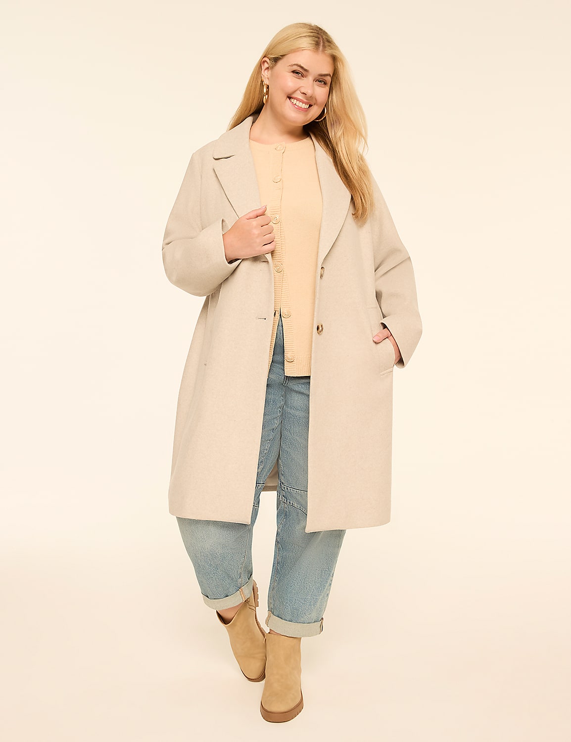 A smiling woman in a beige coat and light blue jeans stands against a plain cream background. Product Image 1