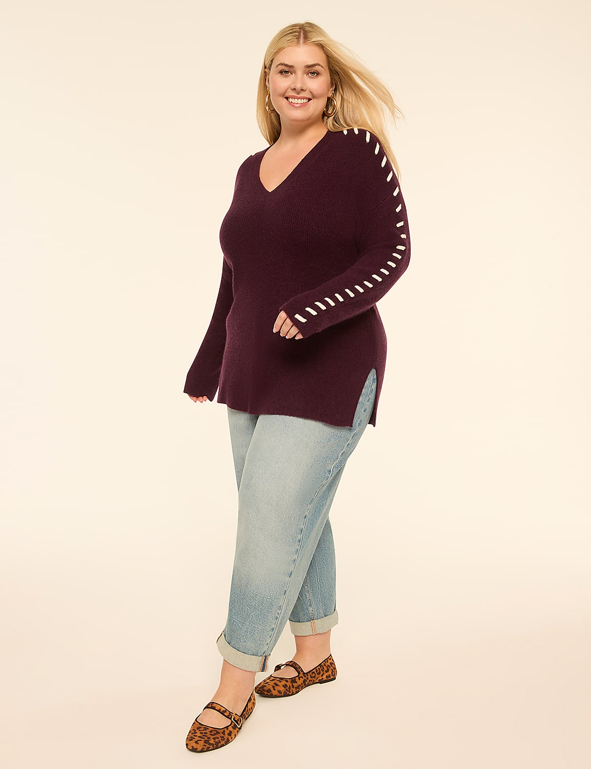A smiling woman in a plum v-neck sweater and light wash jeans stands against a plain cream background. Product Image 3