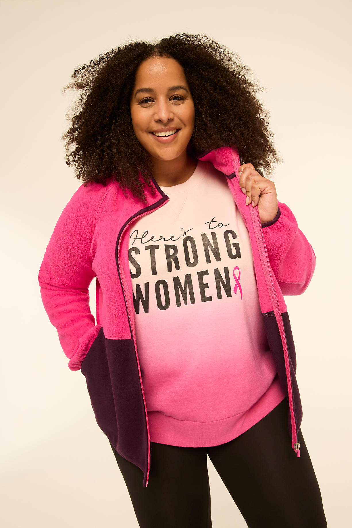A smiling woman with curly hair in the foreground wears a pink and purple zip-up jacket over a white t-shirt that says "HERE'S TO STRONG WOMEN" with a pink ribbon, against a plain, light-colored background. Product Image 6