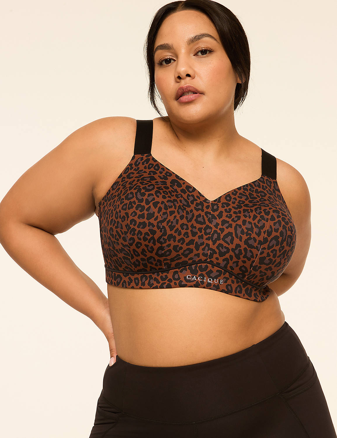 A plus-size woman with dark hair and brown eyes poses in a leopard-print sports bra and black leggings against a plain, light-colored background. Product Image 1