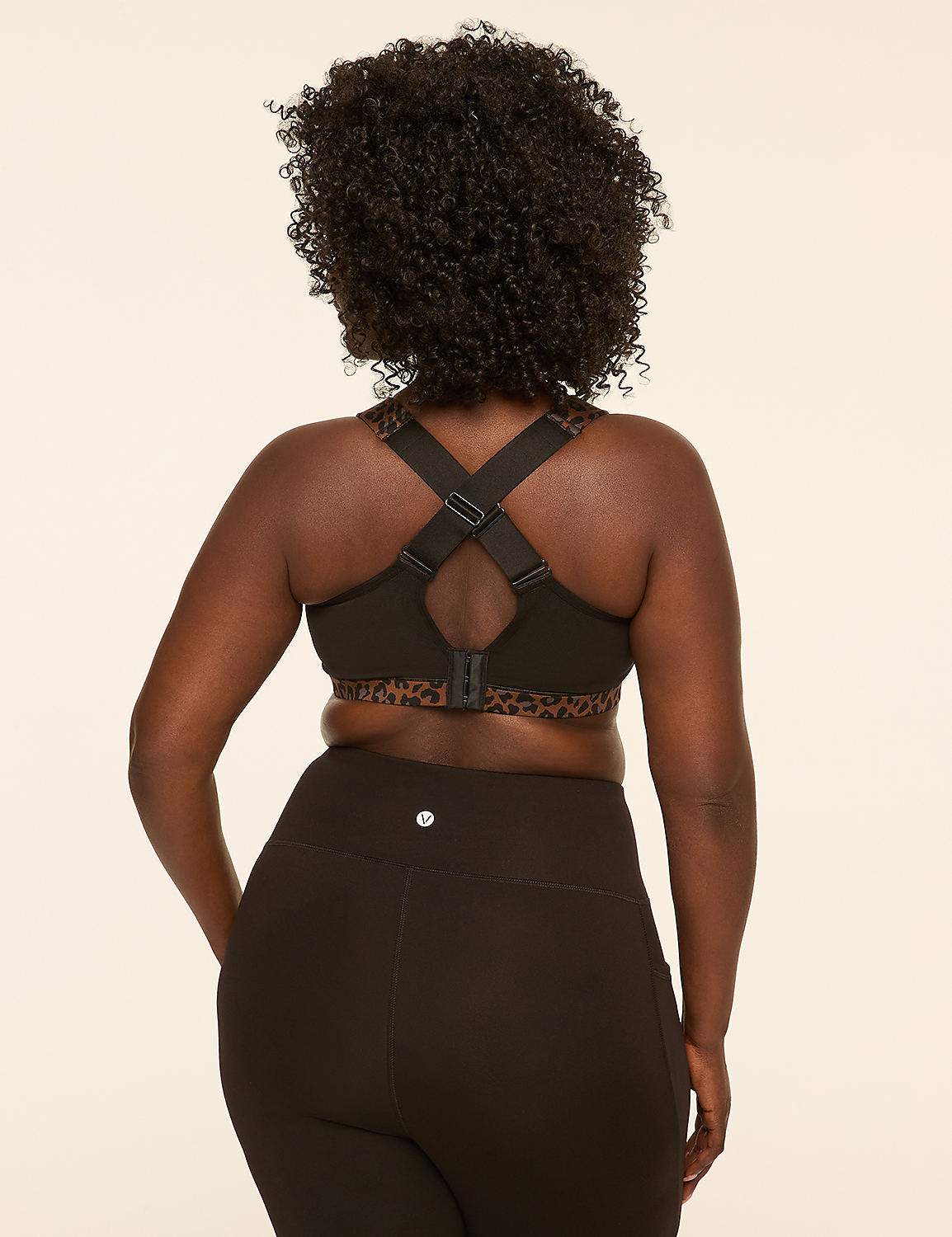 A woman with dark curly hair, wearing a black sports bra with leopard print trim and black leggings, stands against a plain cream background. Product Image 4