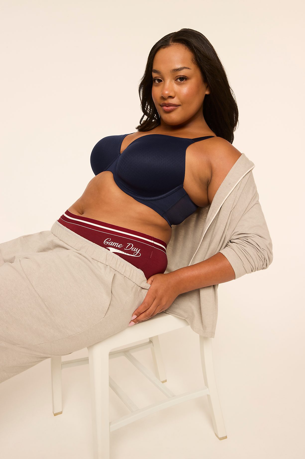 A woman in a navy bra and maroon underwear with "Game Day" written on them, wearing a light-colored zip-up hoodie and beige pants, sits on a white stool against a plain, light background. Product Image 10