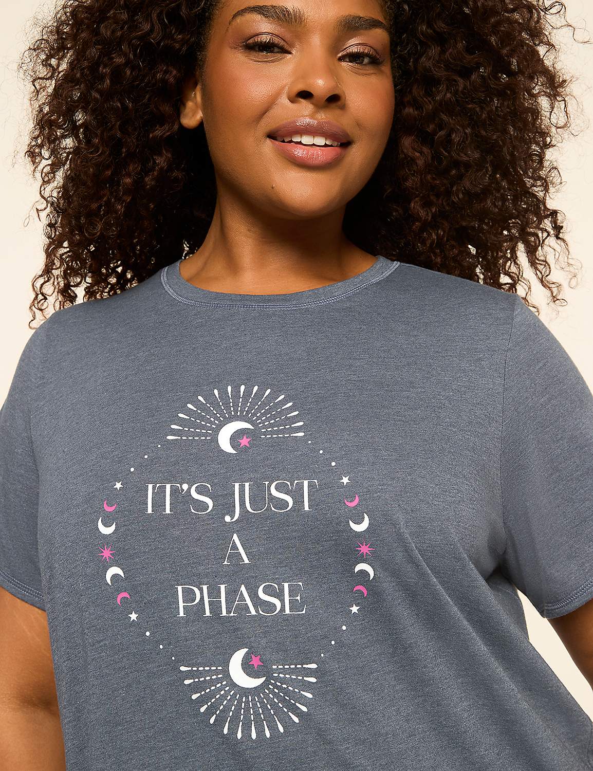 A woman with curly hair smiles as she wears a grey t-shirt with a celestial design and text that reads "IT'S JUST A PHASE" against a plain background. Product Image 3