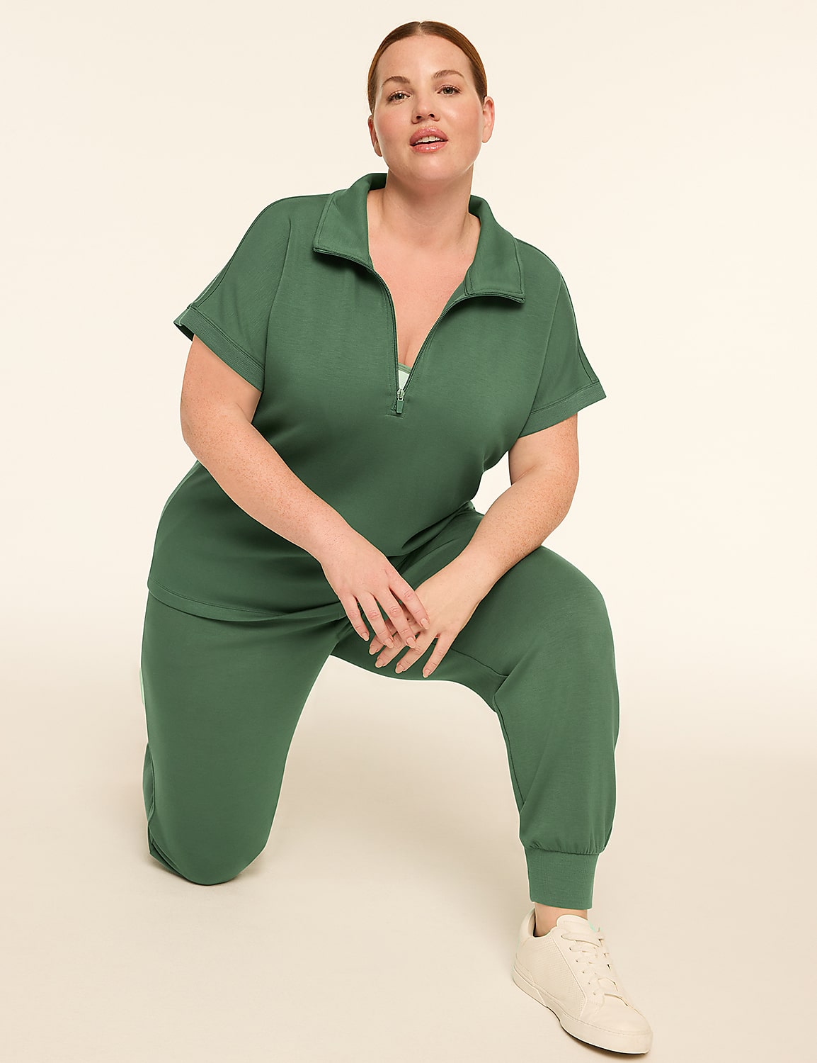 A woman in a green jumpsuit kneels on a light beige floor with a plain, light beige background. Product Image 3