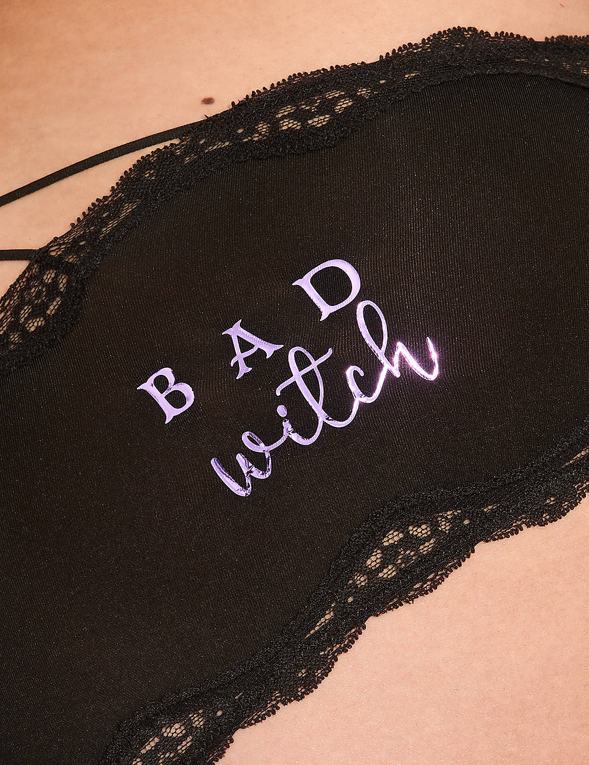 A woman in black lace-trimmed underwear with "bad witch" embroidered on the back is posed against a plain, light-colored wall with soft shadows. Product Image 4