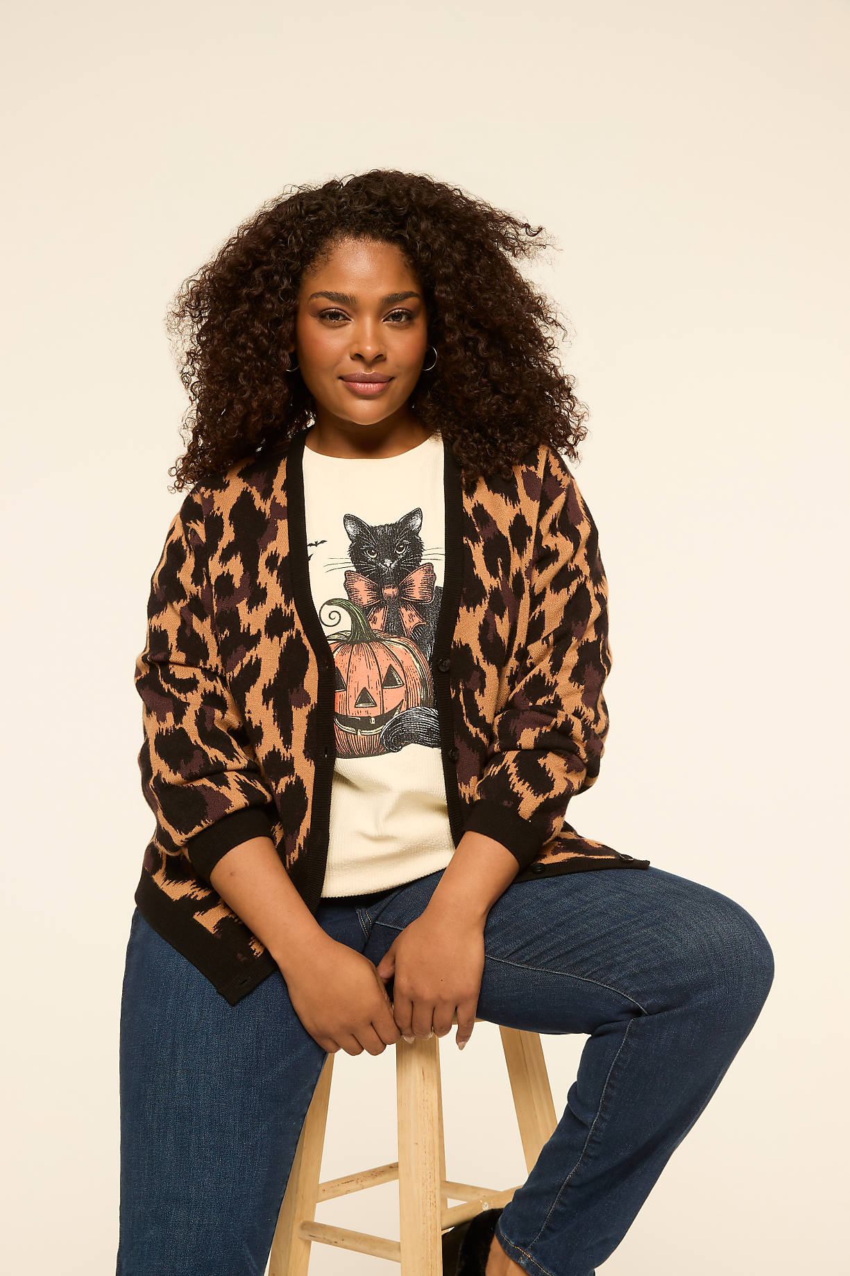 A woman with curly hair sits on a stool in the foreground, wearing a leopard print cardigan over a t-shirt with a black cat and pumpkin graphic, against a plain, light-colored background. Product Image 5