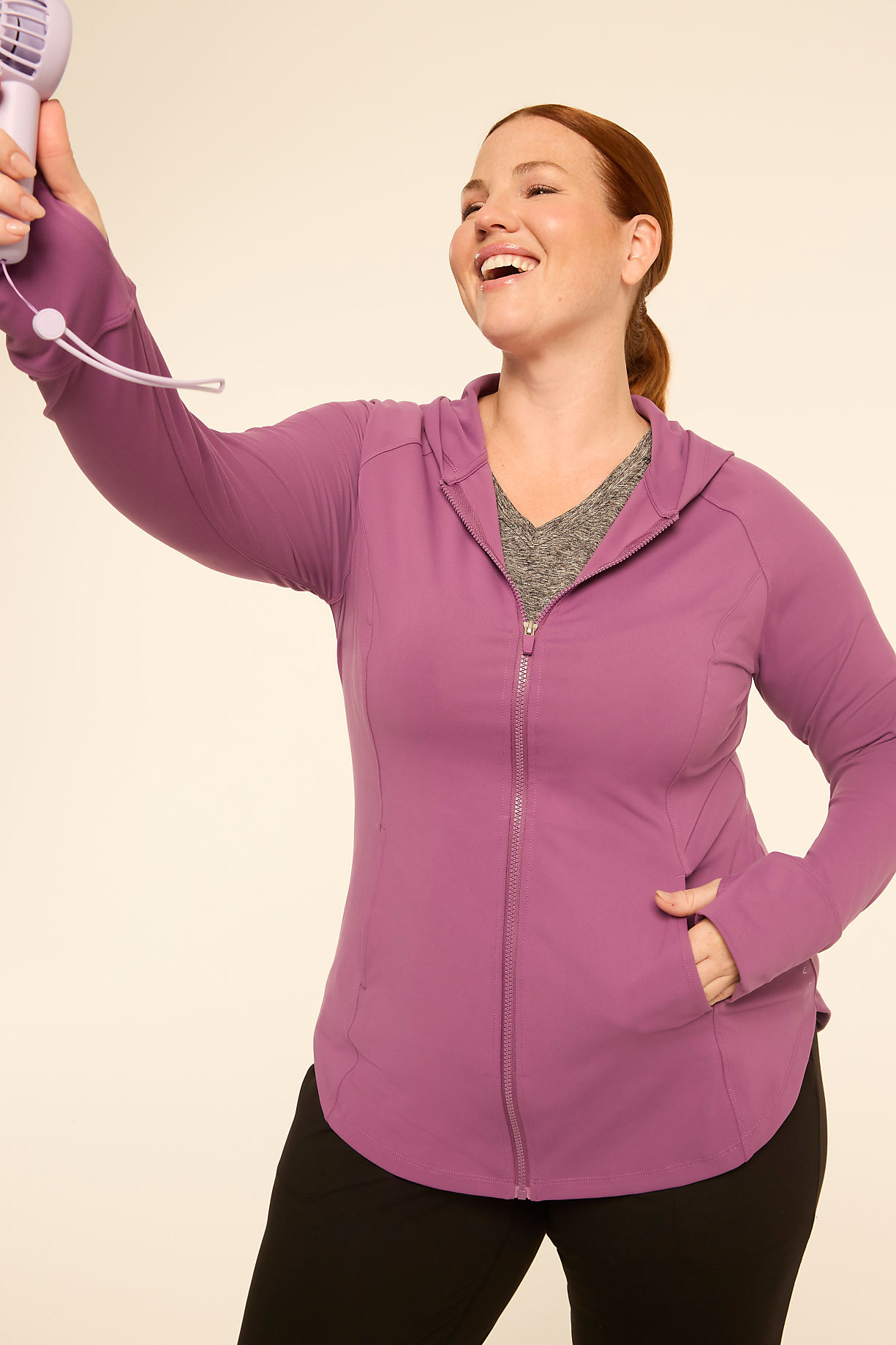 A woman in a purple hoodie and black leggings holds a small fan in her outstretched hand against a plain, light-colored background. Product Image 7