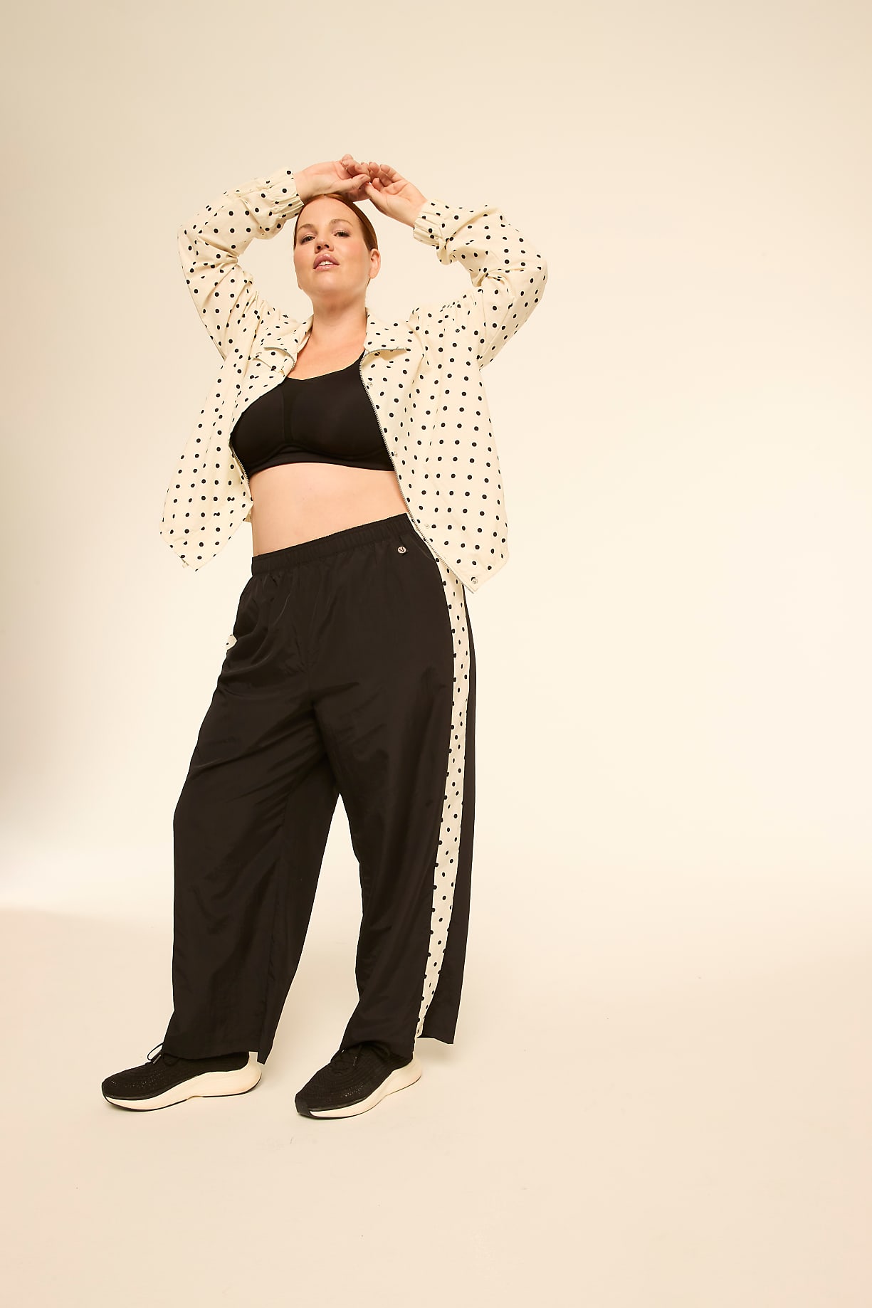 A woman in a black sports bra, a polka-dot shirt, and black track pants stands with her hands behind her head against a plain, light-colored background. Product Image 4