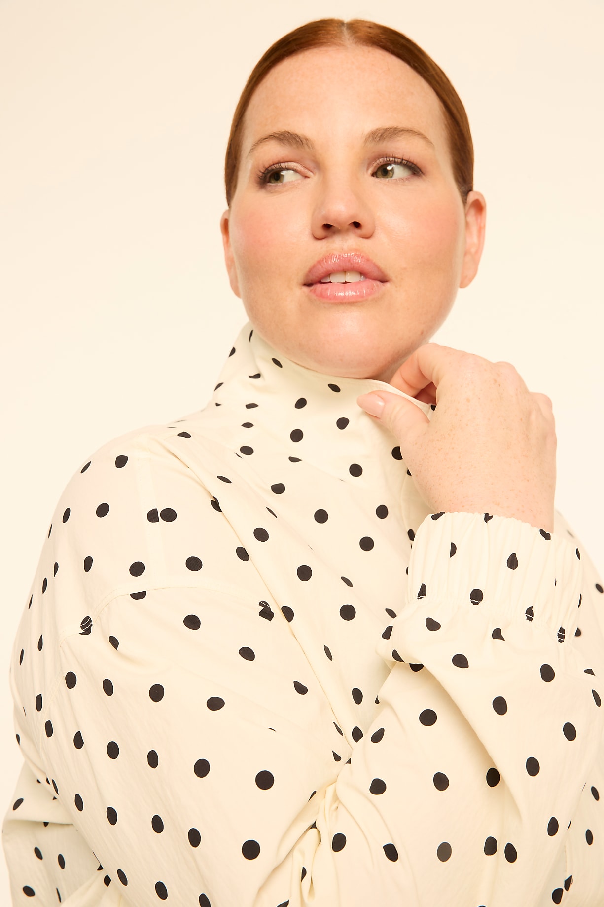 A woman with freckles and rosy cheeks, wearing a cream-colored polka dot blouse, looks to the side against a plain, light background. Product Image 7