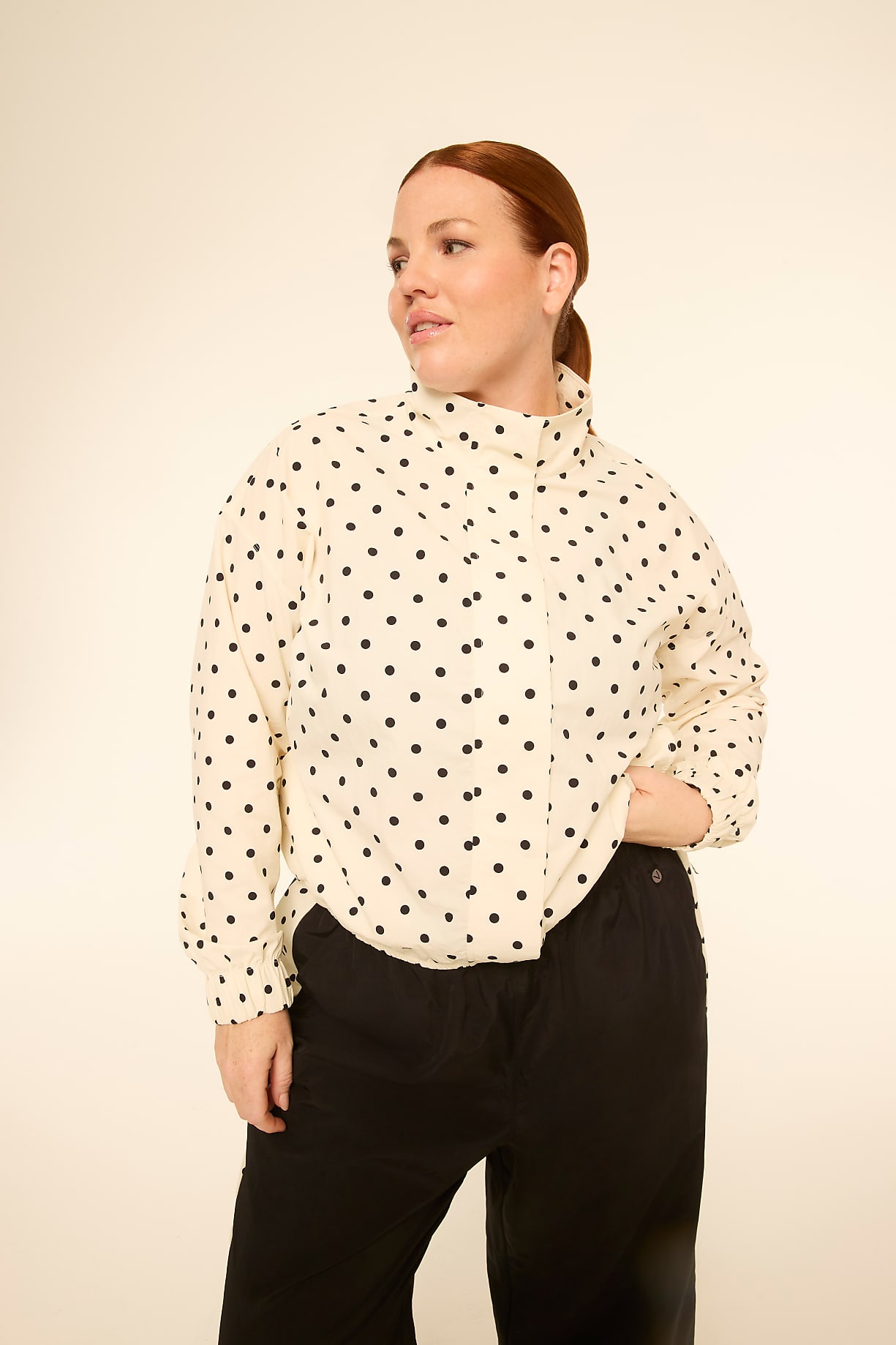A woman with red hair stands in the foreground wearing a cream-colored polka dot blouse and black pants, against a plain, light-colored background. Product Image 8