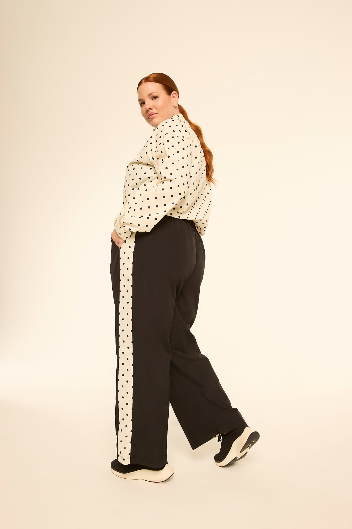 A woman with red hair, wearing a polka dot shirt and black pants with a white stripe, poses against a plain, light-colored background. Product Image 9