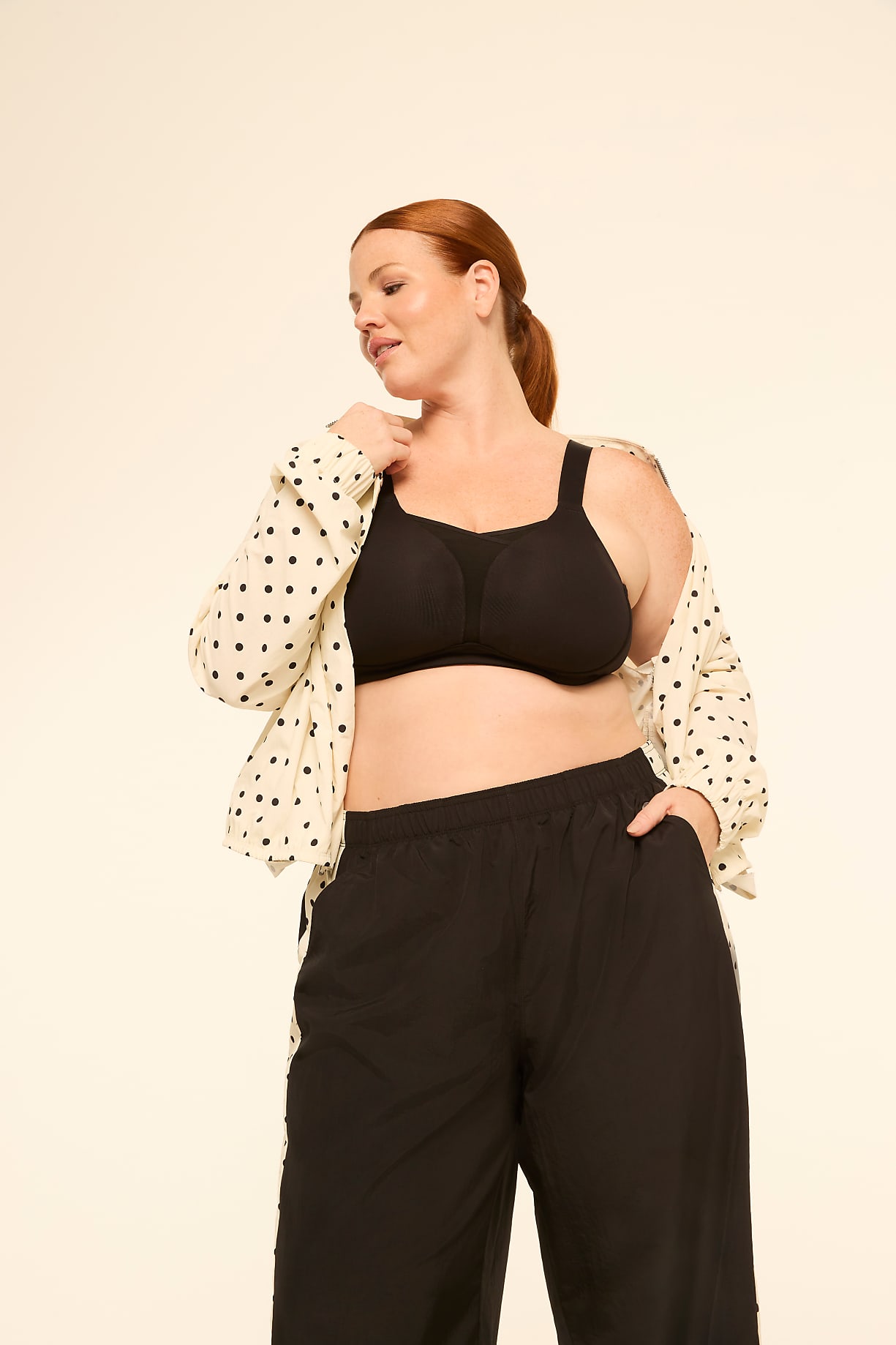 A woman with red hair in a black bra and black pants, with a polka dot shirt draped over her shoulders, stands against a plain cream background. Product Image 10