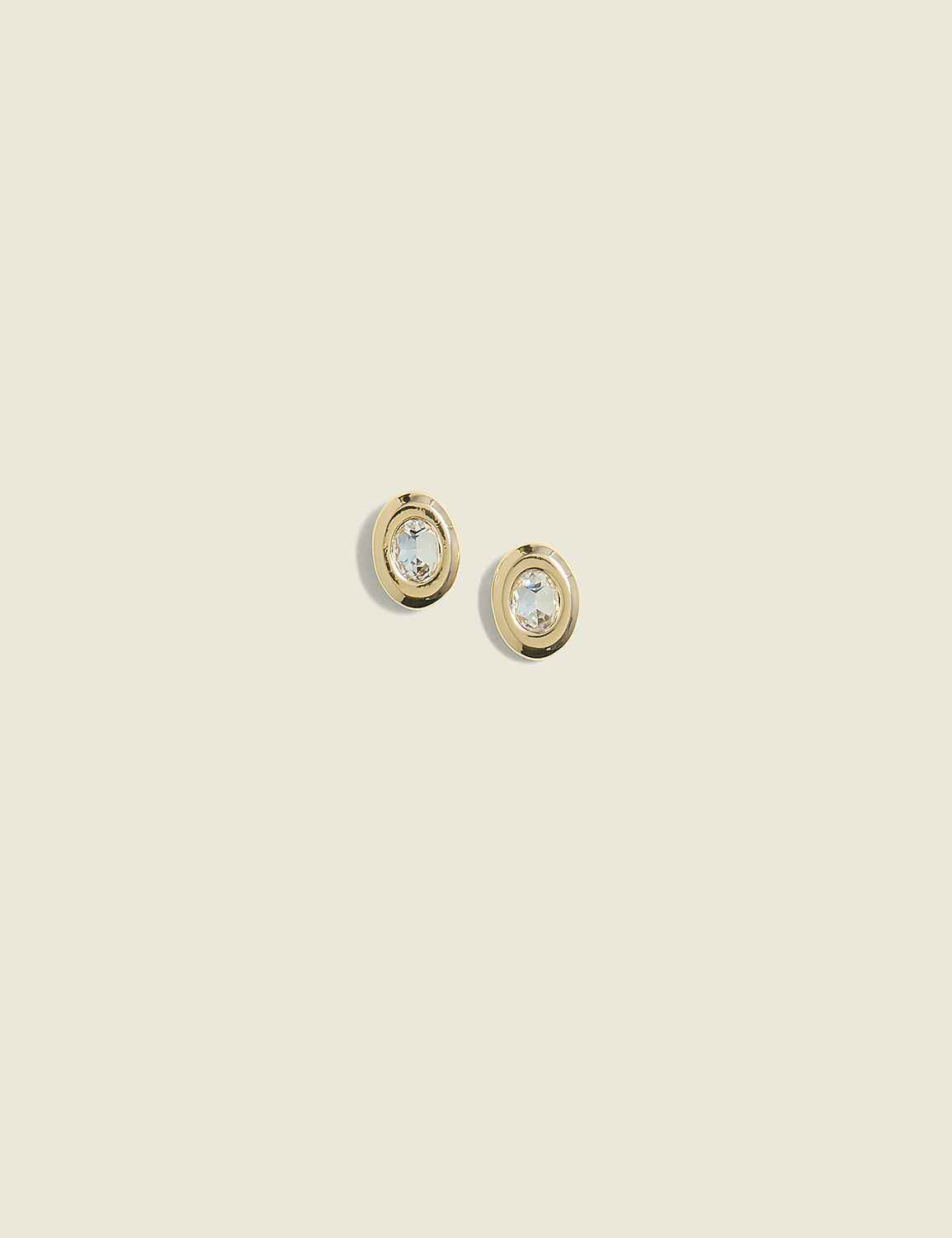 A pair of gold oval earrings with clear stones are centered against a soft, pale yellow background. Product Image 1