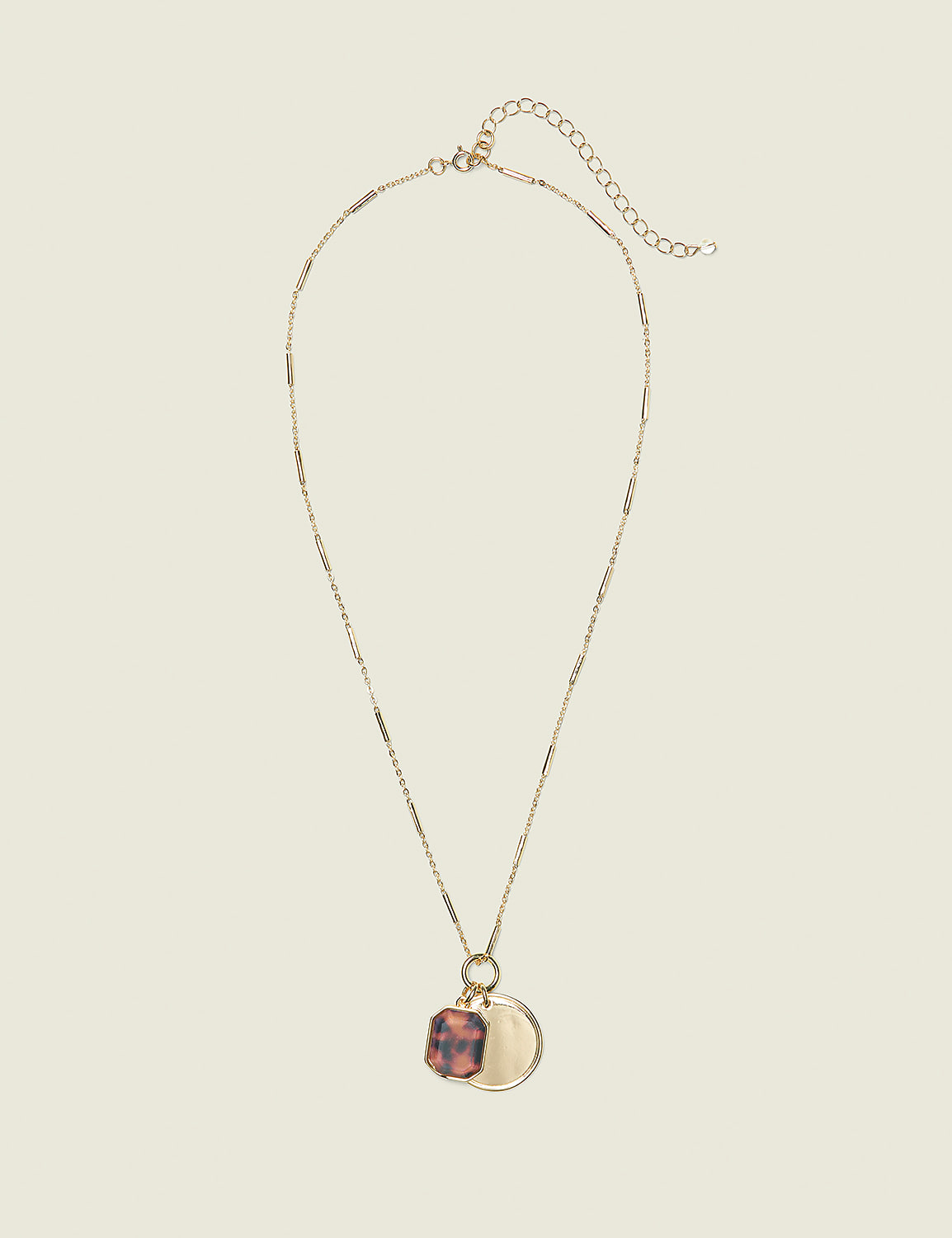 A gold necklace with a tortoise shell charm and a circular pendant hangs against a plain, light beige background. Product Image 1