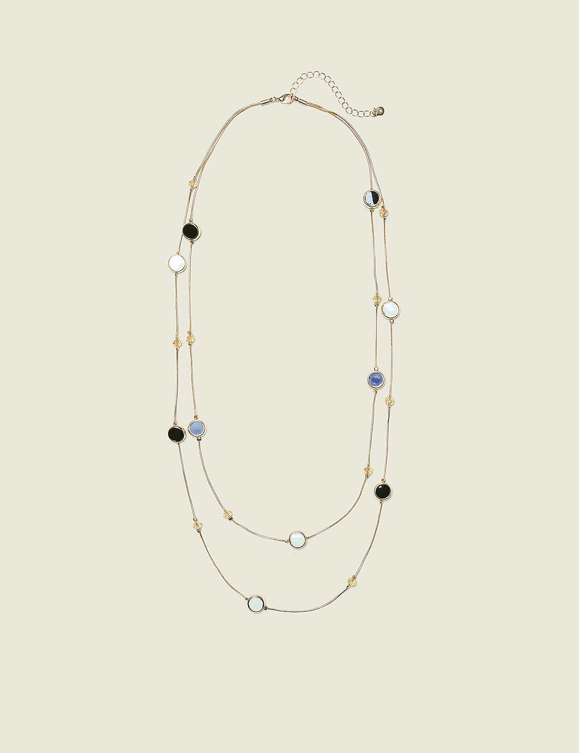 A delicate gold necklace with alternating black, white, and blue circular pendants and small amber beads is laid out against a plain, light beige background. Product Image 1