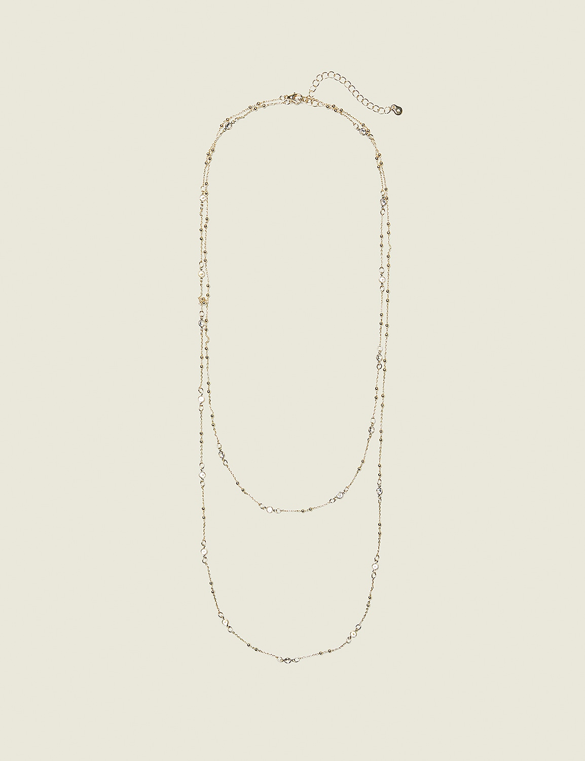 A delicate gold necklace with small charms is laid out against a plain, light beige background. Product Image 1