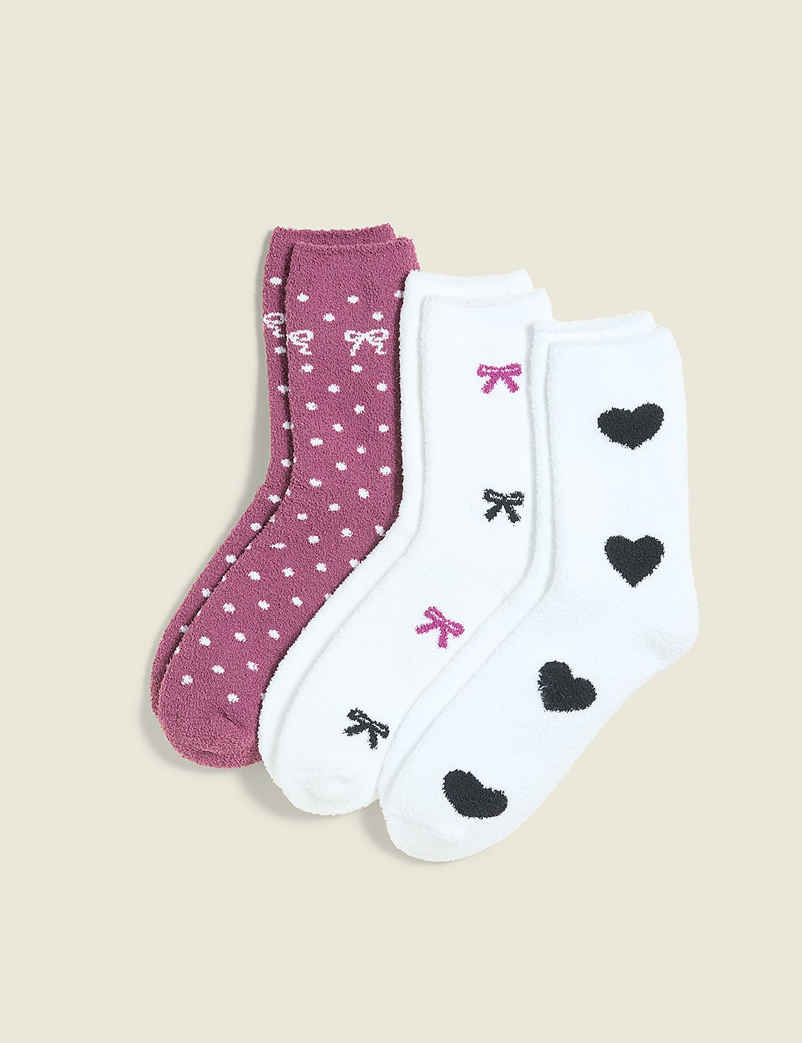 Three pairs of fuzzy socks, one with white polka dots and two with heart and bow patterns, are arranged on a plain, light-colored background. Product Image 1