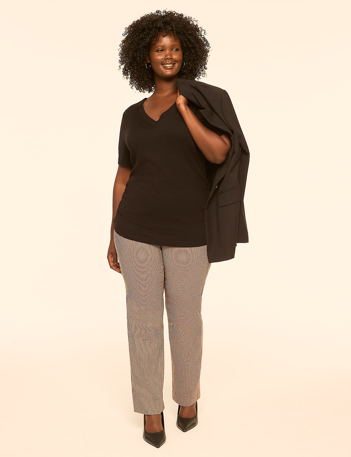 A smiling woman in a black v-neck shirt and checkered pants stands against a plain, light-colored background with a blazer draped over her shoulder. Product Image 3