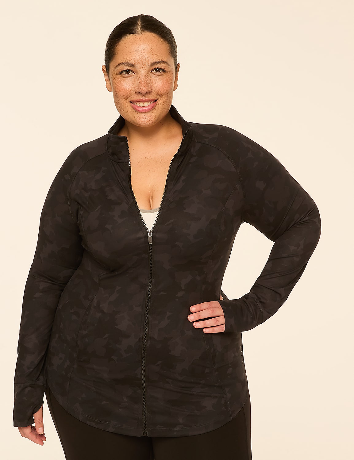 A smiling woman with freckles in a dark camouflage zip-up jacket stands against a plain, light-colored background. Product Image 1