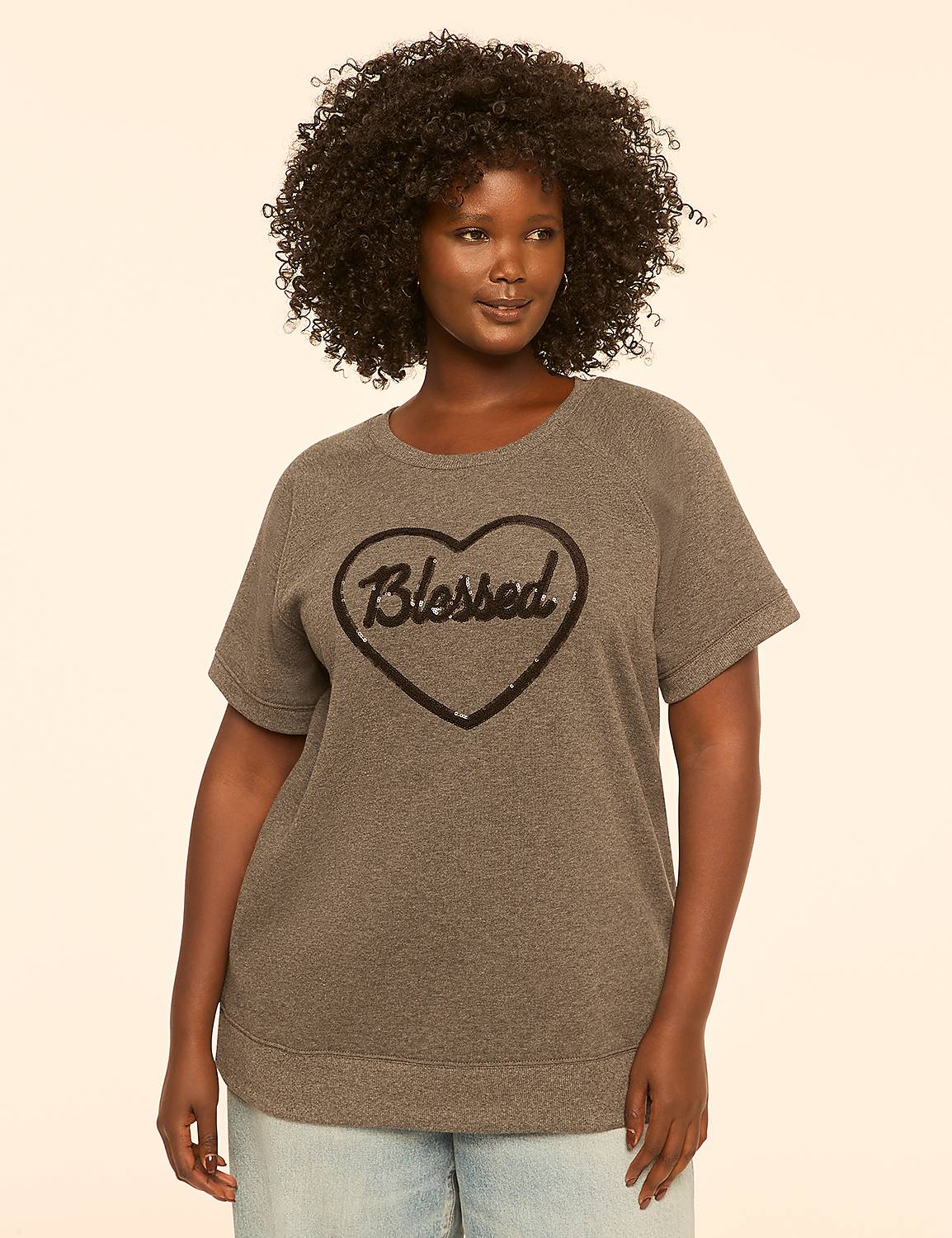 A woman with curly hair stands in the foreground wearing a gray sweatshirt with a sequined heart that says "Blessed," against a plain, light-colored background. Product Image 1