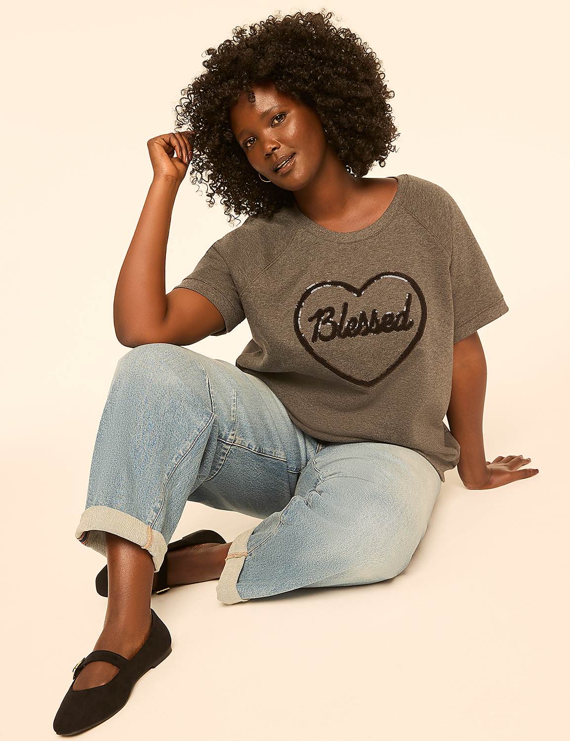 A woman with curly hair sits on a plain background, wearing a grey t-shirt with a sequined heart that says "Blessed" and light-wash cuffed jeans. Product Image 3