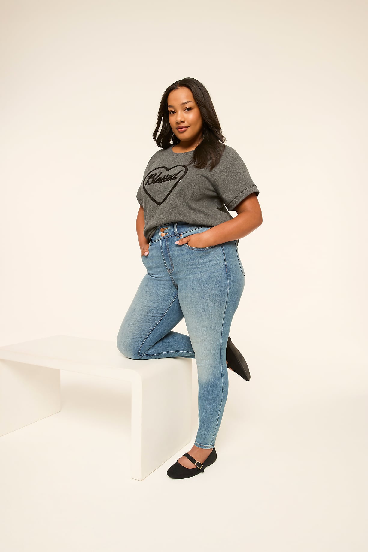A woman in a grey t-shirt and blue jeans stands with one leg resting on a white bench against a plain white background. Product Image 5
