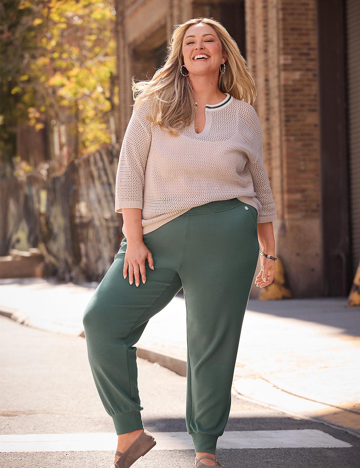 A smiling woman in a beige sweater and green joggers stands on a street with a brick building in the background. Product Image 4