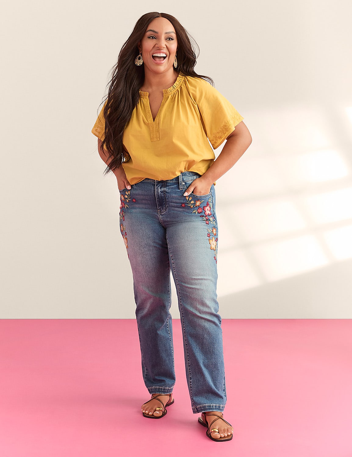 A smiling woman in a yellow top and embroidered jeans stands on a pink floor against a light background with shadows. Product Image 5