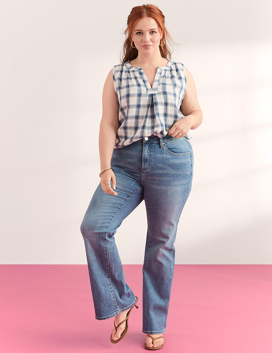 A woman with auburn hair stands in a plaid top and flared jeans against a pink and white background. Product Image 4