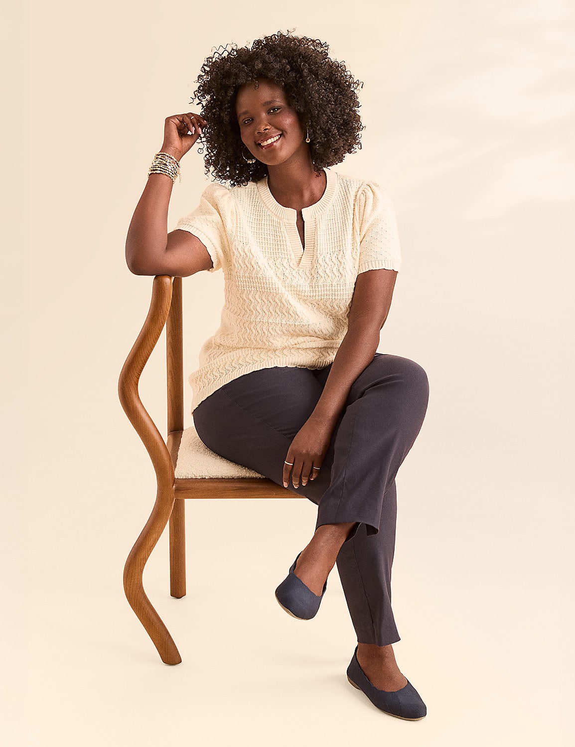 A smiling woman with dark curly hair sits on a wooden chair with a light-colored seat, her legs crossed, against a plain, light background. Product Image 4