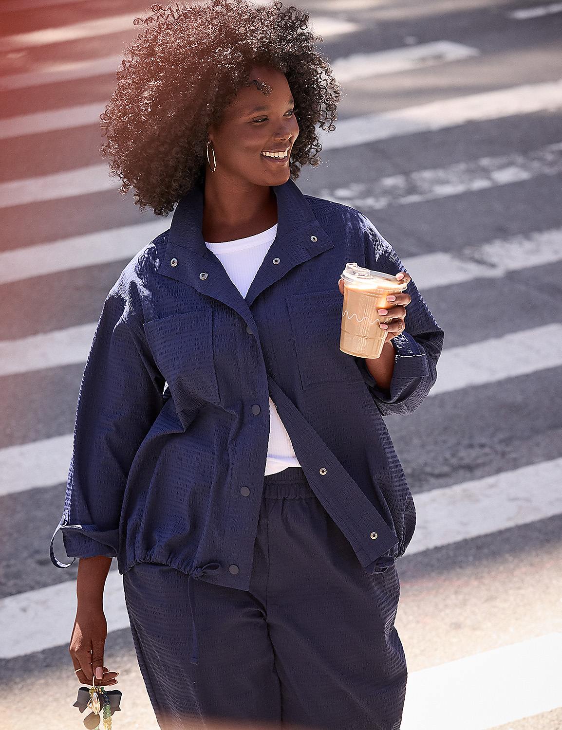 A smiling woman in a navy blue jacket and pants walks across a crosswalk holding a coffee cup and keys. Product Image 4