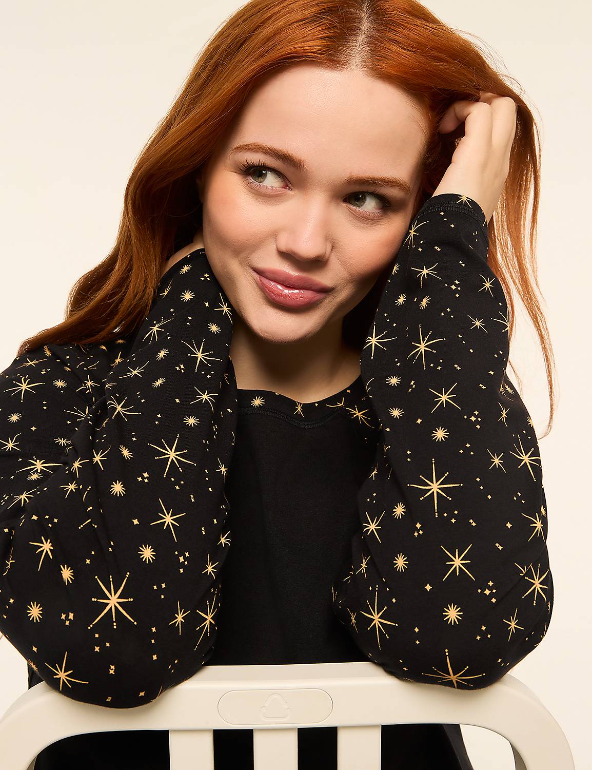 A young woman with red hair and a star-patterned top rests her chin on a white chair against a plain background. Product Image 4