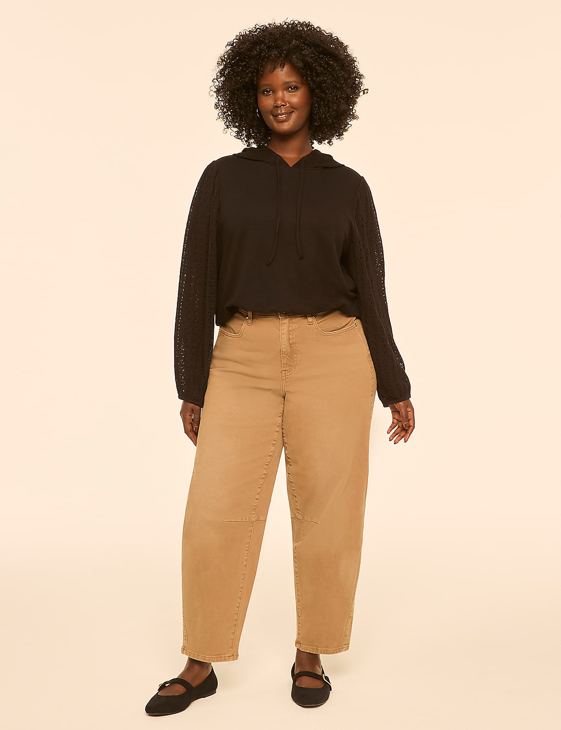 A woman with dark, curly hair stands in the foreground wearing a black hoodie and tan pants, against a plain, light-colored background. Product Image 3